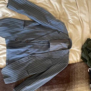 Randig blå kavaj från Yves Saint Laurent - Snygg blå kavaj från Yves Saint Laurent med tunna vertikala ränder i olika nyanser av blått och grått. Klassisk passform med två knappar framtill och fickor. Lätt och luftig känsla, perfekt för dig som vill sticka ut med stil.