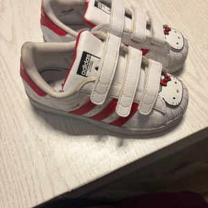 Adidas Superstar med Hello Kitty-design - Säljer ett par vita Adidas Superstar med röd Hello Kitty-design. Skorna har kardborreband och de klassiska tre ränderna i rött. Perfekta för den som älskar unika sneakers med en twist!