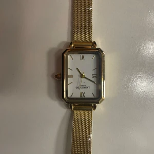 Guldfärgad klocka från Lorezo - Elegant guldfärgad klocka från Lorezo med rektangulär urtavla och romerska siffror. Klockan har ett mesh-armband i guld och en vit urtavla som ger en stilren look. Perfekt accessoar för att lyfta din stil.
