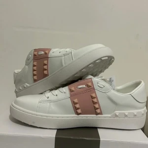 Valentino Garavani Rockstud sneakers - Snygga vita sneakers från Valentino Garavani med rosa detaljer och ikoniska nitar. Skorna har en stilren design med snörning och en bekväm sula. Perfekta för att ge din outfit en lyxig touch.