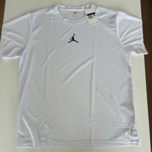 Vit t-shirt från Jordan - Säljer en vit t-shirt från Jordan med det ikoniska Jumpman-logotypen på bröstet. T-shirten är tillverkad i ett lätt och andningsbart syntetmaterial, perfekt för träning eller vardagsbruk. Den har en normal passform och korta ärmar. Passa på att köpa denna stilrena t-shirt! 🏀