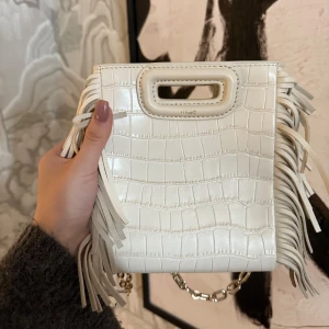 Vit axelväska med fransar från Maje - Snygg vit axelväska från Maje med krokodilpräglat mönster och fransar på sidorna. Väskan har en praktisk kedjerem och ett handtag upptill. Perfekt för att ge din outfit en trendig touch! Den lilla storleken och aldrig använd