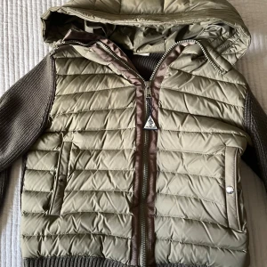 Beige och brun jacka från Moncler - Säljer en snygg beige och brun jacka från Moncler. Jackan har en quiltad design med dragkedja och huva. Ärmarna är ribbade och ger en cool kontrast till det quiltade materialet. Perfekt för kyligare dagar!