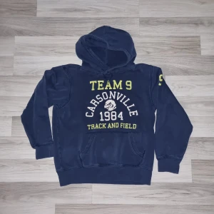 Mörkblå hoodie från H&M  - Säljer en cool mörkblå hoodie från H&M med tryck 'TEAM 9 CARSONVILLE 1984 TRACK AND FIELD' i gult och vitt. Perfekt för en sportig look med känguruficka och huva. Passar perfekt för vardagsäventyr!