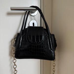Svart axelväska med kedjedetaljer - Snygg svart axelväska i krokodilmönstrat syntetmaterial. Väskan har en stilren design med en kedja som axelrem och dubbla handtag. Perfekt för att ge din outfit en trendig touch.