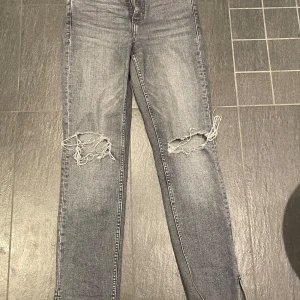 Grå jeans med slitningar - Snygga grå jeans med slitningar på knäna. De har en klassisk femficksdesign och en dragkedja framtill. Perfekta för en avslappnad stil.