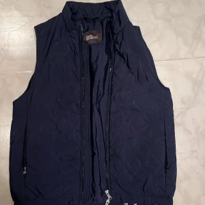Mörkblå quiltad väst från Oscar Jacobson - Snygg mörkblå väst från Oscar Jacobson med quiltat mönster och dragkedja framtill.  Visar sömmar har brustit.