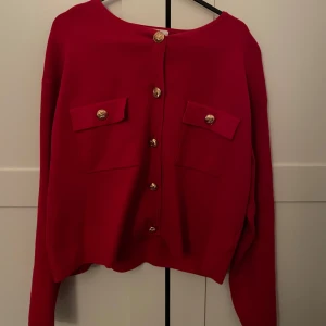 Röd kofta från H&M - Säljer en snygg röd kofta från H&M med guldknappar och två dekorativa fickor framtill. Perfekt för att ge en färgklick till din outfit. Koftan har långa ärmar och en klassisk rund halsringning.