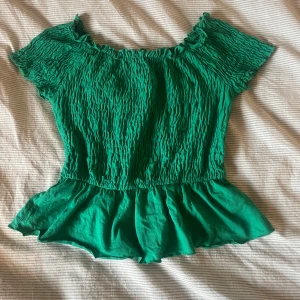 Grön topp från Gina Tricot - Säljer en grön topp från Gina Tricot med korta ärmar och volangdetaljer. Toppen har en smockad design upptill och en lösare passform nedtill. Perfekt för en somrig look! 🌿