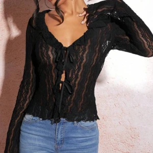 Svart spetsblus med knyt - Snygg svart spetsblus med långa ärmar och knyt framtill. Perfekt för en stilfull look med en touch av elegans. Blusen har en v-ringad design och volangdetaljer längs kanterna.
