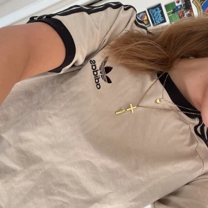 Beige t-shirt från Adidas - Snygg beige t-shirt från Adidas med klassiska svarta ränder på ärmarna och svart krage samt Adidas-logga på bröstet.