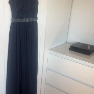 Mörkblå långklänning med spetsdetaljer - Elegant mörkblå långklänning med vackra spetsdetaljer på överdelen och ett glittrande bälte i midjan. Perfekt för en formell tillställning. Använd ett tillfälle. Hund finns i hemmet. 