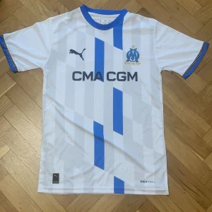 Vit och blå fotbollströja från Puma - Säljer en snygg vit fotbollströja från Puma med blå detaljer och korta ärmar. Tröjan har ett randigt mönster och klubbmärket på bröstet. Perfekt för fotbollsfans som vill visa sitt stöd.