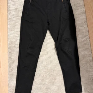 Svarta byxor med dragkedja - Snygga svarta byxor med dragkedjor vid fickorna. De har en högmidjad design och en slim passform som ger en stilren look. Perfekta för en chic och modern stil.