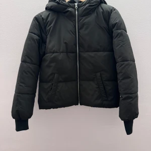 Svart pufferjacka med huva - Säljer en svart pufferjacka med huva och dragkedja framtill. Jackan har långa ärmar och är perfekt för kyligare dagar. Den quiltade designen ger extra värme och stil.