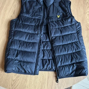 Svart dunväst från Lyle & Scott - Snygg svart dunväst från Lyle & Scott med dragkedja och quiltad design. Perfekt för lager-på-lager under kyliga dagar. Den har en diskret logga på bröstet och är lätt att matcha med olika outfits.