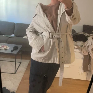 Beige trenchcoat med bälte -  beige trenchcoat med bälte i midjan och knappar framtill. Jackan har långa ärmar och en klassisk krage.  för vår och höst!