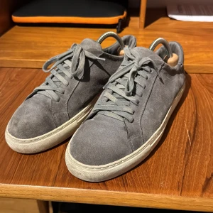 Grå sneakers från Axel Arigato - Snygga grå sneakers i mocka från Axel Arigato med klassisk snörning och vit sula. Perfekta för en stilren look.