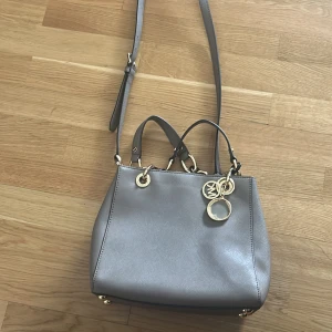 Grå axelväska från Michael Kors - Snygg grå axelväska från Michael Kors med gulddetaljer och justerbar axelrem. Väskan har en dragkedja upptill och dekorativa ringar. Perfekt för att bära med stil!
