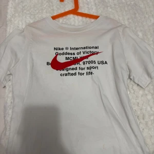 Vit t-shirt från Nike - Säljer en vit t-shirt från Nike med tryck i svart och rött på framsidan. T-shirten har korta ärmar och är perfekt för en sportig look. Den är tillverkad i ett mjukt och bekvämt material. Använd fåtal gånger 