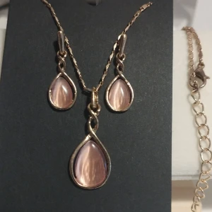 Guldhalsband och örhängen med rosa stenar - Elegant smyckesset med ett halsband och matchande örhängen. Båda har droppformade rosa stenar infattade i en guldfärgad ram. Perfekt för att ge en sofistikerad touch till din outfit. Halsbandet är 22 cm långt utan hänge samt 25 cm med hänge. Det finns också en förlängning på 5 cm. Obs: Ej äkta guld!