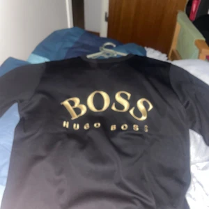 Svart tröja från Hugo Boss - Säljer en stilren svart tröja från Hugo Boss med guldtryck på framsidan. Tröjan har en klassisk passform och är perfekt för en avslappnad men ändå elegant look.