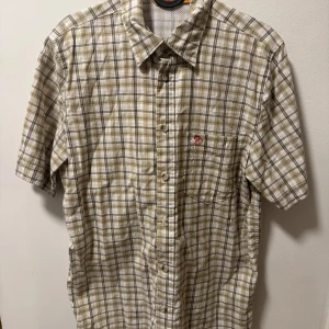 Rutig kortärmad skjorta från Fjällräven - Säljer en stilren rutig skjorta från Fjällräven i beige och grå toner. Skjortan har korta ärmar och en klassisk krage med knappar framtill. Perfekt för en avslappnad stil. Skriv gärna om du har frågor eller vill ha mer bilder. 
