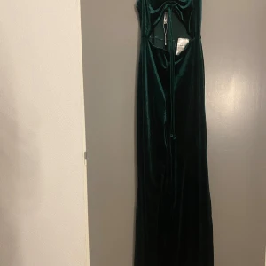 Mörkgrön långklänning från FB Sister - Elegant mörkgrön långklänning från FB Sister i sammet. Klänningen har en öppen rygg och knytning framtill, vilket ger en stilfull look. Perfekt för speciella tillfällen.