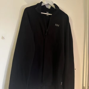 Svart fleecejacka från Helly Hansen - Säljer en svart fleecejacka från Helly Hansen med dragkedja framtill. Rätt lång, passar Unisex
