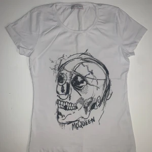 Vit t-shirt från Alexander McQueen - Säljer en vit t-shirt från Alexander McQueen med ett coolt tryck av en skalle på framsidan. T-shirten är tillverkad i 95% bomull och 5% polyester, vilket gör den mjuk och bekväm. Perfekt för dig som vill ha en unik stil! Fint skick, strl S  Nypris ca 2000 SEK