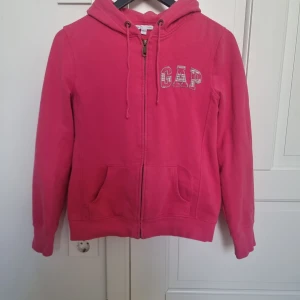 Rosa hoodie från GAP - Säljer en snygg rosa hoodie från GAP med dragkedja och luva. Den har ett broderat GAP-märke på bröstet och är väl omhändertagen. Finns lite korta lösa trådar från användning.