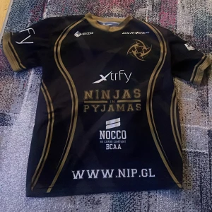 Svart och guld Ninjas in Pyjamas tröja - Säljer en svart och guld tröja från Ninjas in Pyjamas med flera sponsorloggor som EIZO, xtrfy och NOCCO. Tröjan har korta ärmar och en rund halsringning. Perfekt för fans av e-sportlaget! 🎮 2015 edition😍