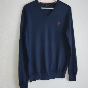 Mörkblå tröja från Fred Perry - Säljer en stilren mörkblå tröja från Fred Perry med v-ringning och broderad logga i orange på bröstet. Tröjan har långa ärmar och ribbade muddar vid ärmslut och nederkant. Perfekt för en klassisk look.