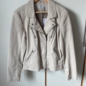 Beige bikerjacka från ONLY - Ljusbeige jacka i fuskskinn från ONLY med dragkedjedetaljer och quiltade axlar. Klassisk bikerstil med asymmetrisk dragkedja och flera fickor. Insvängd. Ny med prislappen kvar. Längd 63 cm. 