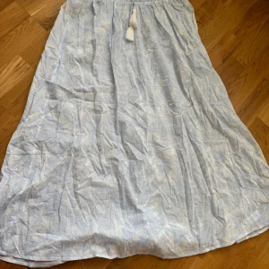 Ljusblå långkjol med mönster - Säljer en ljusblå långkjol med subtilt mönster och elastisk midja. Kjolen har en dekorativ tofsdetalj framtill och är perfekt för varma dagar.