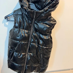 Svart glansig dunväst - Snygg svart glansig dunväst med dragkedja framtill. Perfekt för att hålla värmen med stil. Västen har en hög krage och är quiltad för extra komfort. Ett måste för garderoben!