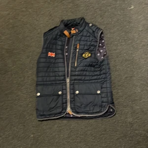 Mörkblå quiltad väst med broderad logga - Snygg mörkblå quiltad väst med dragkedja och broderad logga på bröstet. Västen har även en brittisk flagga som detalj. Perfekt för kyliga dagar.