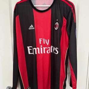 AC Milan fotbollströja med Ronaldinho 80 - Säljer en klassisk AC Milan fotbollströja från Adidas med Ronaldinho och nummer 80 på ryggen. Tröjan är randig i rött och svart med långa ärmar och Fly Emirates som sponsorlogga på framsidan. Perfekt för samlare eller fans av laget!