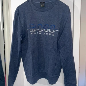 Blå tröja från Hugo Boss - Säljer en stilren blå tröja från Hugo Boss med broderad logga i vitt och blått på bröstet. Tröjan har en rund halsringning och är slim fit. Perfekt för en avslappnad men ändå snygg look.