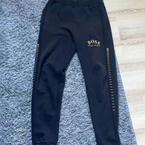 Svarta mjukisbyxor från Hugo Boss - Snygga svarta mjukisbyxor från Hugo Boss med gulddetaljer längs sidorna och broderad logga på framsidan. Byxorna har en elastisk midja och en bakficka. Perfekta för en avslappnad stil.