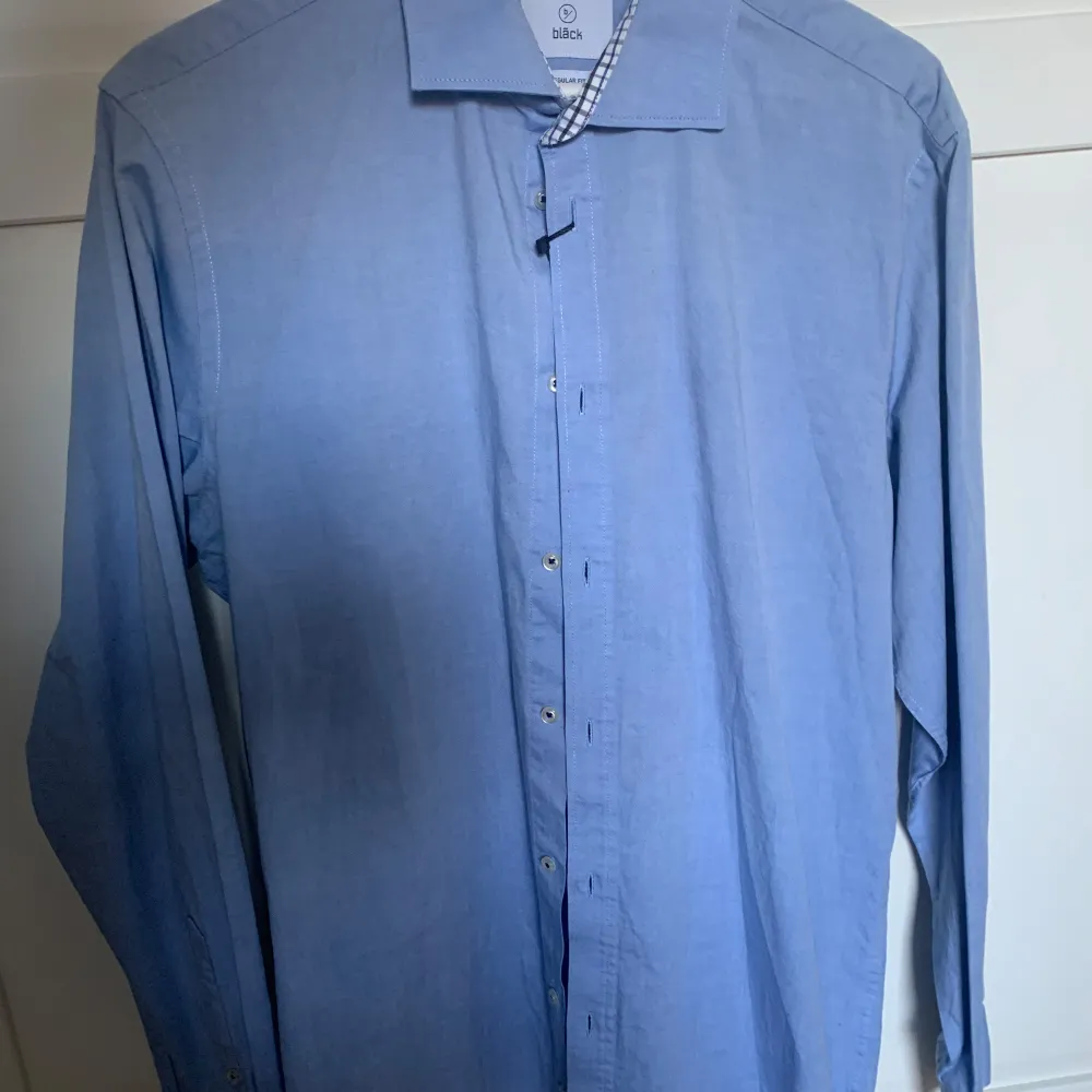 Snygg blå skjorta från Bläck med klassisk krage och knappar framtill. Skjortan har ett diskret rutigt mönster på insidan av kragen och är perfekt för en stilren look.. Paidat.