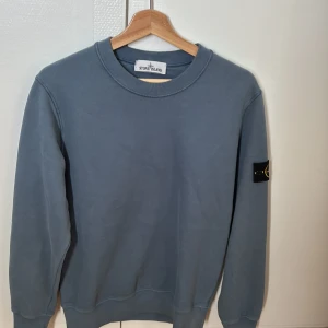 Stone Island tröja - Säljer en stilren blå sweatshirt från Stone Island. Tröjan har långa ärmar och en rund halsringning. Perfekt för en avslappnad stil med en touch av lyx. Märkeslogga på ärmen ger en extra detalj.
