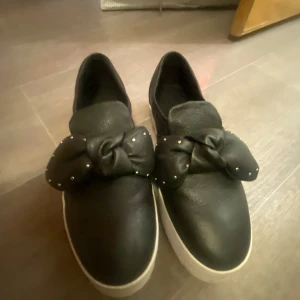 Svarta sneakers med rosett - Snygga svarta sneakers med en stor rosett framtill och små nitar som detaljer. Skorna har en vit sula som ger en fin kontrast. Perfekta för en trendig look. Kostade 700kr men säljer för 200 