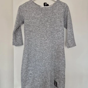 Grå stickad klänning med knytband - Mysig grå stickad klänning med trekvartsärmar och ett stilrent knytband i ryggen. Perfekt för kyligare dagar med sin mjuka och bekväma passform. En liten etikettdetalj finns längst ner på klänningen.