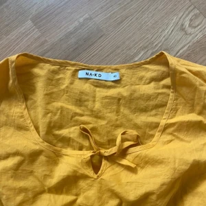 Gul blus från NA-KD - Säljer en solgul blus från NA-KD med korta ärmar och en söt knytdetalj vid halsen. Blusen har en smockad midja som ger en fin passform och volangdetaljer vid ärmslut och nederkant. Perfekt för vår och sommar! Aldrig använd