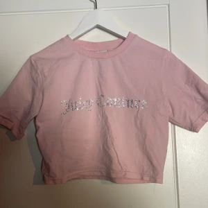 Rosa croppad t-shirt från Juicy Couture - Säljer en söt rosa croppad t-shirt från Juicy Couture med glittrig logga på framsidan. Perfekt för en avslappnad stil med en touch av glamour. T-shirten har korta ärmar och rund halsringning.