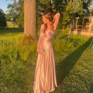 Champagnefärgad långklänning i satin - Elegant champagnefärgad långklänning i satin med en vacker glans. Klänningen har en v-ringad design och är ärmlös, vilket ger en stilren och sofistikerad look. Perfekt för speciella tillfällen. Köpt för 3500kr 