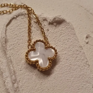 Guldhalsband med fyrklöverberlock - Elegant halsband i guld med en fyrklöverformad berlock. Berlocken har en vit insida och är omgiven av en detaljerad guldkant. Perfekt för att ge en touch av lyx till din stil.