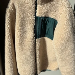 Beige teddyjacka från H&M - Mysig beige teddyjacka från H&M med en grön detalj på bröstet. Jackan har en dragkedja framtill och är perfekt för kyligare dagar. Den är långärmad och har en mjuk och fluffig textur som håller dig varm och bekväm. Passar storlek XS-152 (lite större i storleken)