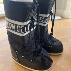 Svarta Moon Boots med snörning - Säljer ett par svarta Moon Boots med snörning och vit logga. Knappast kommit till användning Perfekta för vintern med sin robusta design och varma foder. De har en hög modell som ger extra skydd mot kyla och snö. Passar bra för kalla dagar och snöiga äventyr.